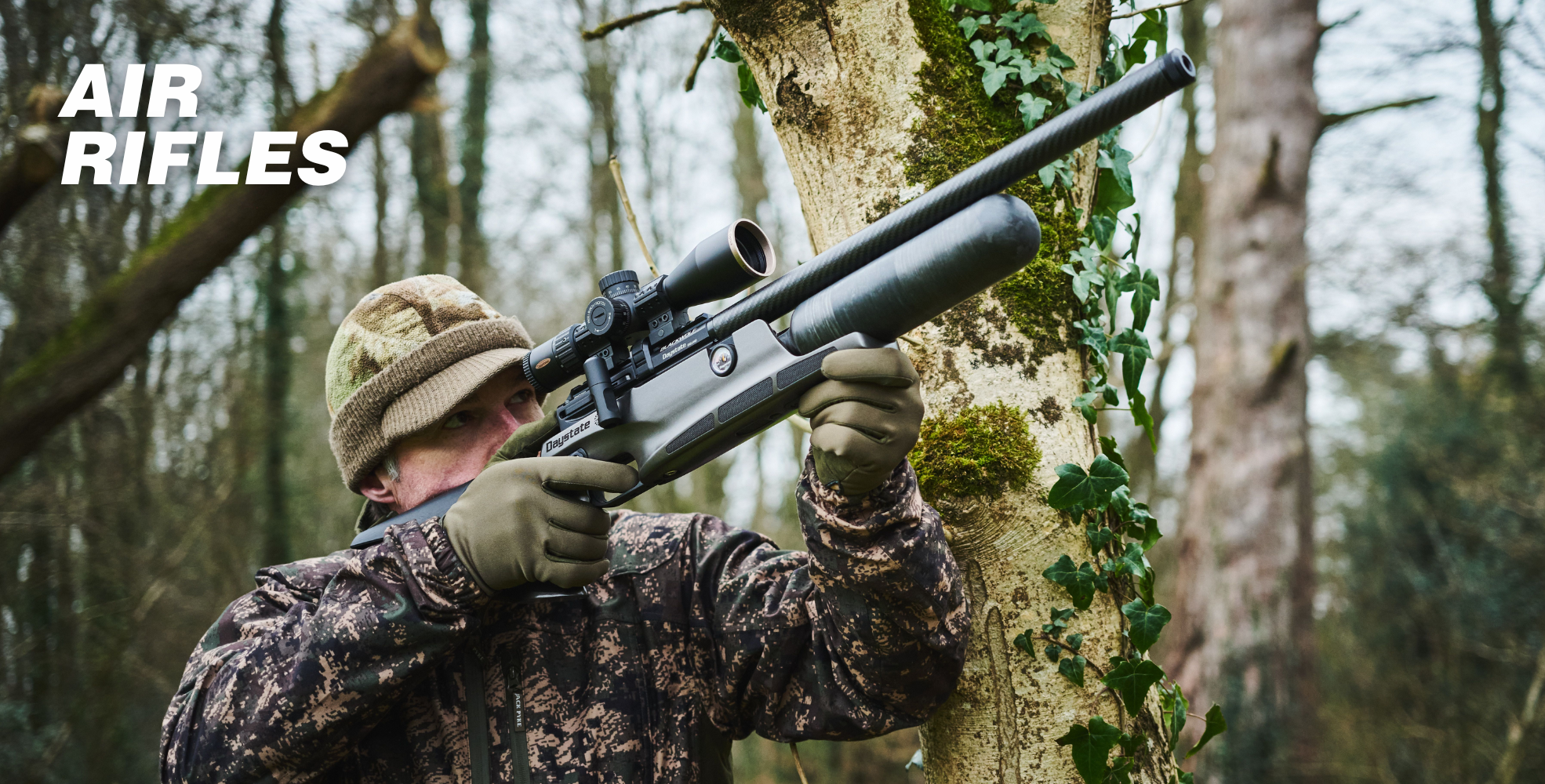 Person in camouflage gear aiming an air rifle while standing next to a tree in a forest. Text on the image reads "AIR RIFLES.