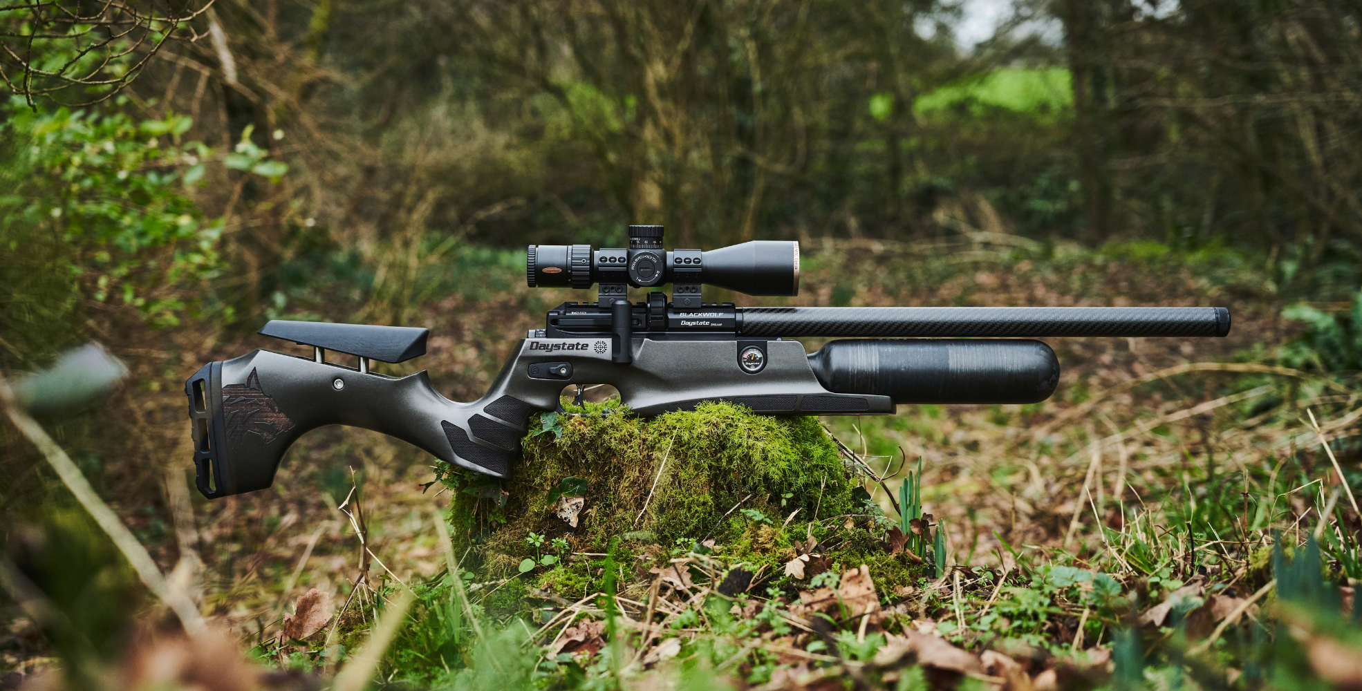 A Daystate Blackwolf air rifle with a mounted scope rests on a mossy rock in a forest setting, surrounded by leaves and trees.