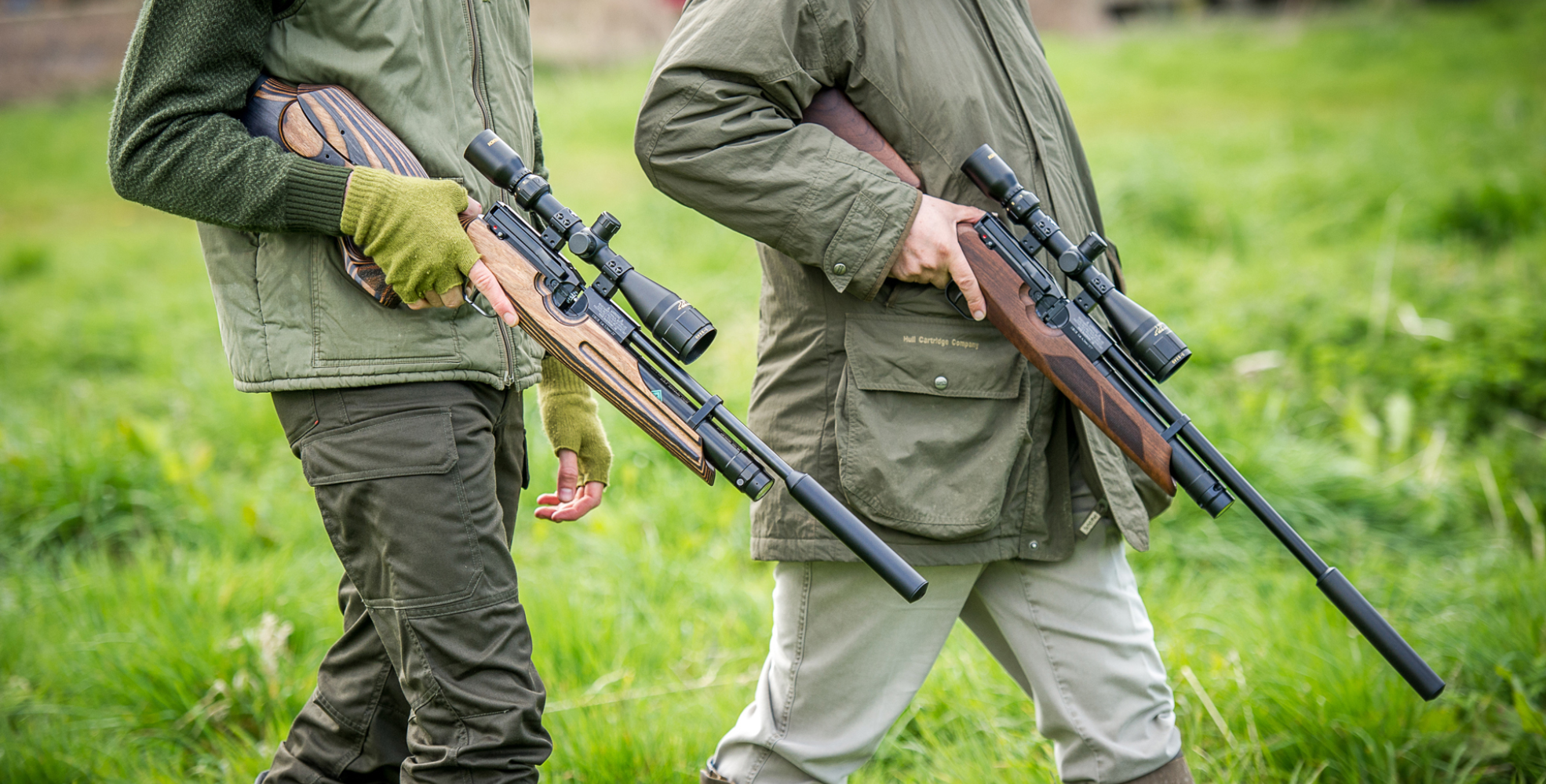 Two people walk on grass, each holding a scoped rifle with a silencer, wearing outdoor jackets and gloves&mdash;perfect for those seeking new shooting locations or following a trusted shooting guide.