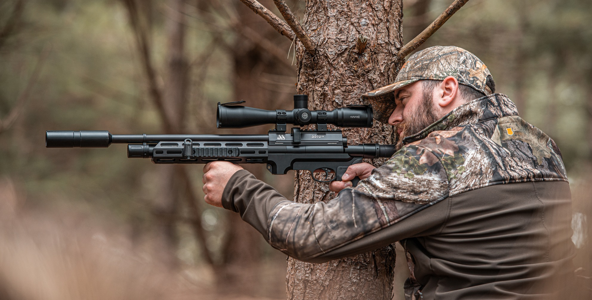 How To Zero Your Scope 1 A man in camouflage clothing aims a scoped rifle, using a tree for support in a wooded outdoor setting, focusing intently on proper rifle sighting.
