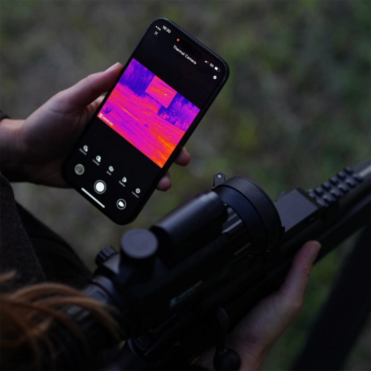 Hikmicro Panther 2.0 PH50L Thermal Imaging LRF Rifle Scope 3 A person holds a smartphone showing a thermal image in one hand and a rifle with a Hikmicro Panther 2.0 PH50L Thermal Imaging LRF Rifle Scope in the other.