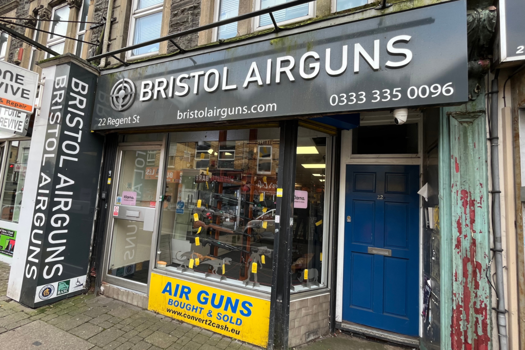 Shopfront of "Bristol Airguns" on Regent Street, featuring a black sign with white lettering and a large window displaying various air guns and accessories. A yellow "AIR GUNS BOUGHT & SOLD" sign highlights customer purchasing trends.