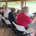 People of various ages sit in a row at an indoor shooting range, aiming rifles at targets outside. Just as last year sales reflected changing buying behaviour, they concentrate on their targets from white chairs under a covered structure.