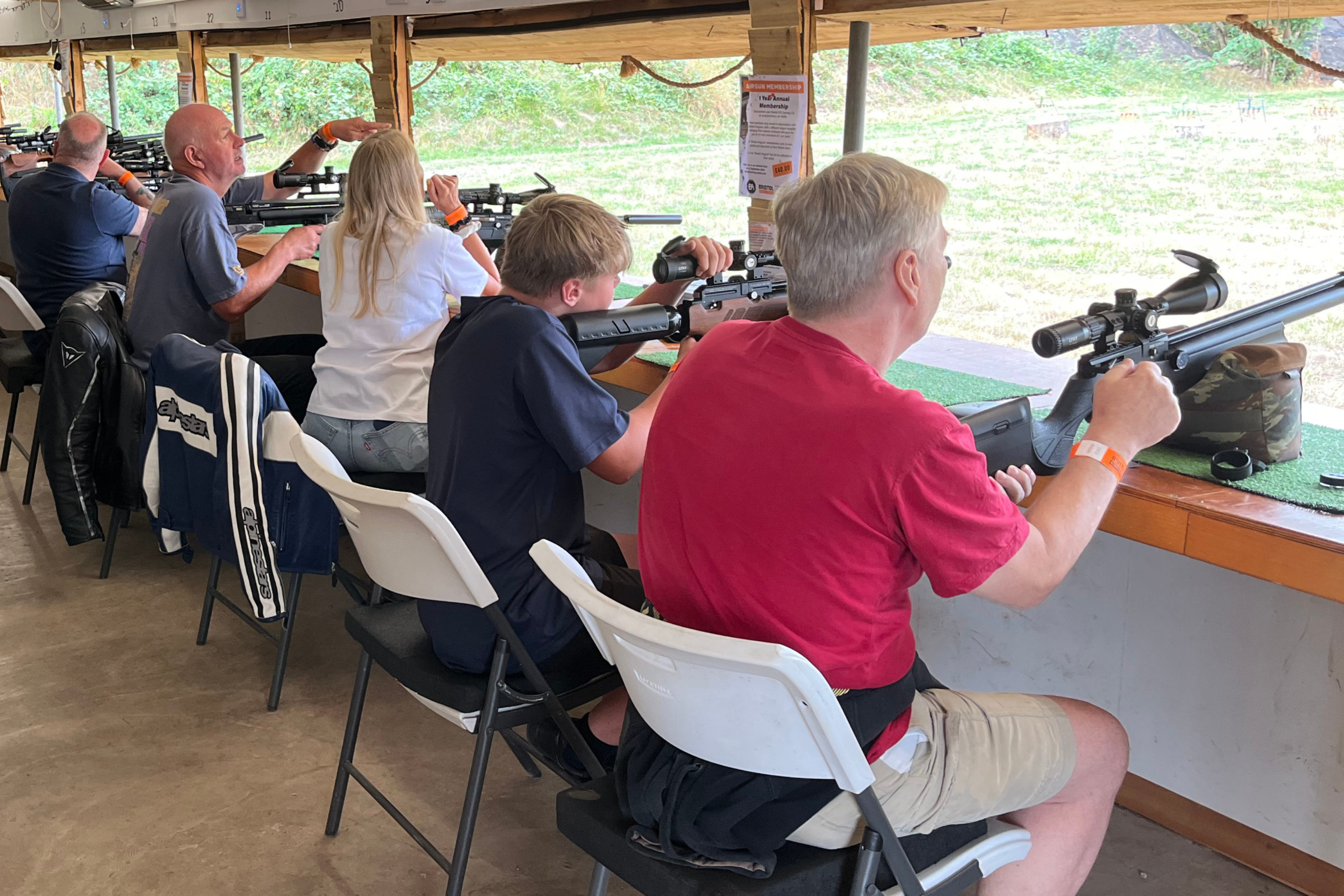 People of various ages sit in a row at an indoor shooting range, aiming rifles at targets outside. Just as last year sales reflected changing buying behaviour, they concentrate on their targets from white chairs under a covered structure.