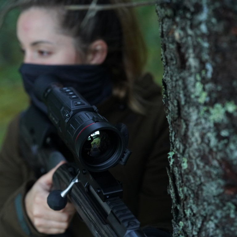HIKMICRO Thunder Pro Zoom 2.0 TQ60Z Optical Thermal Rifle Scope 5 A partial view of a person's face beside a lichen-covered tree trunk, with blurred green foliage and a HIKMICRO Thunder Pro Zoom 2.0 TQ60Z Optical Thermal Rifle Scope subtly visible in the background.